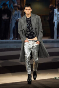 Male model on a runway wearing a long gray textured coat over a black floral top and shiny silver pants, with black ankle boots.