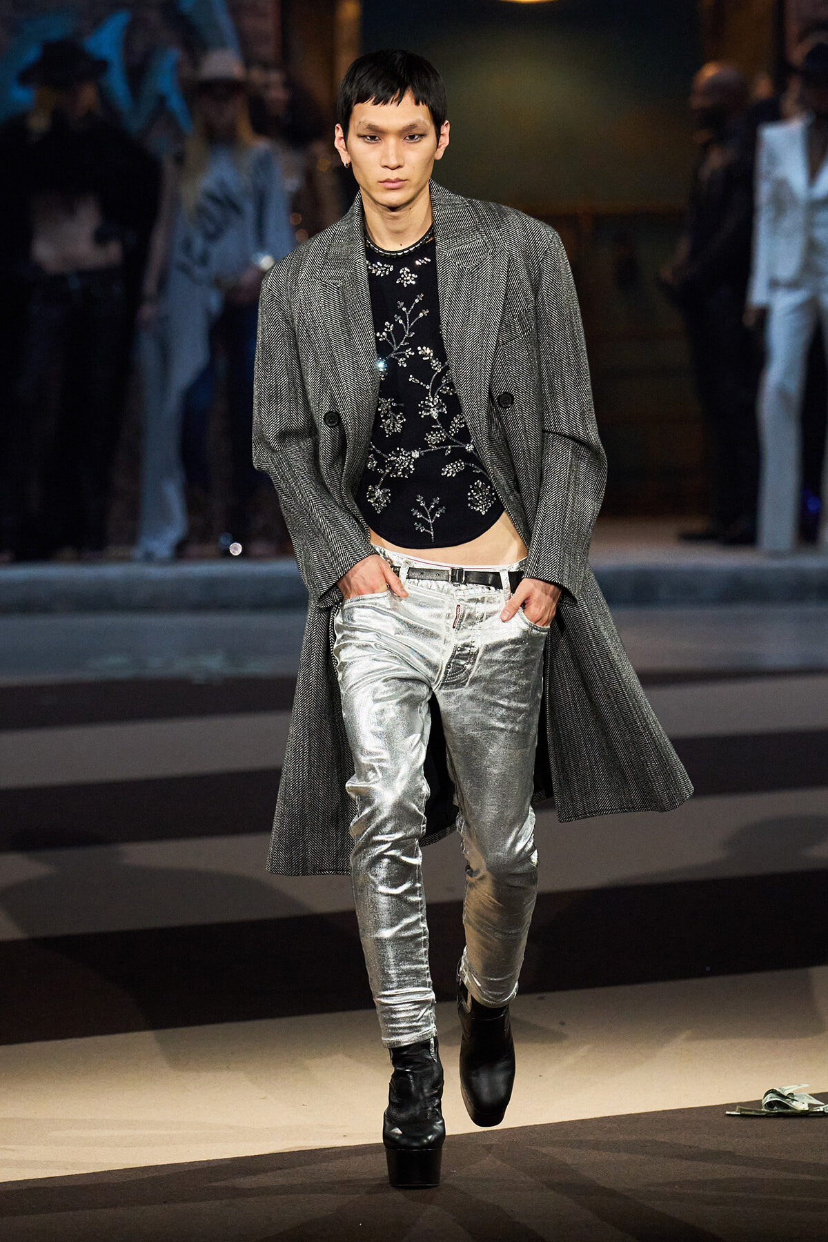 Male model on a runway wearing a long gray textured coat over a black floral top and shiny silver pants, with black ankle boots.