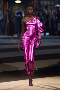 Model walking a fashion runway in a shiny magenta one-shoulder jumpsuit with a large puff sleeve, matching glove on one arm, and strappy heels.