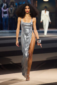 Model walking the runway in a silver metallic gown with a high slit and strappy heels, bold red lips, and voluminous curly hair.