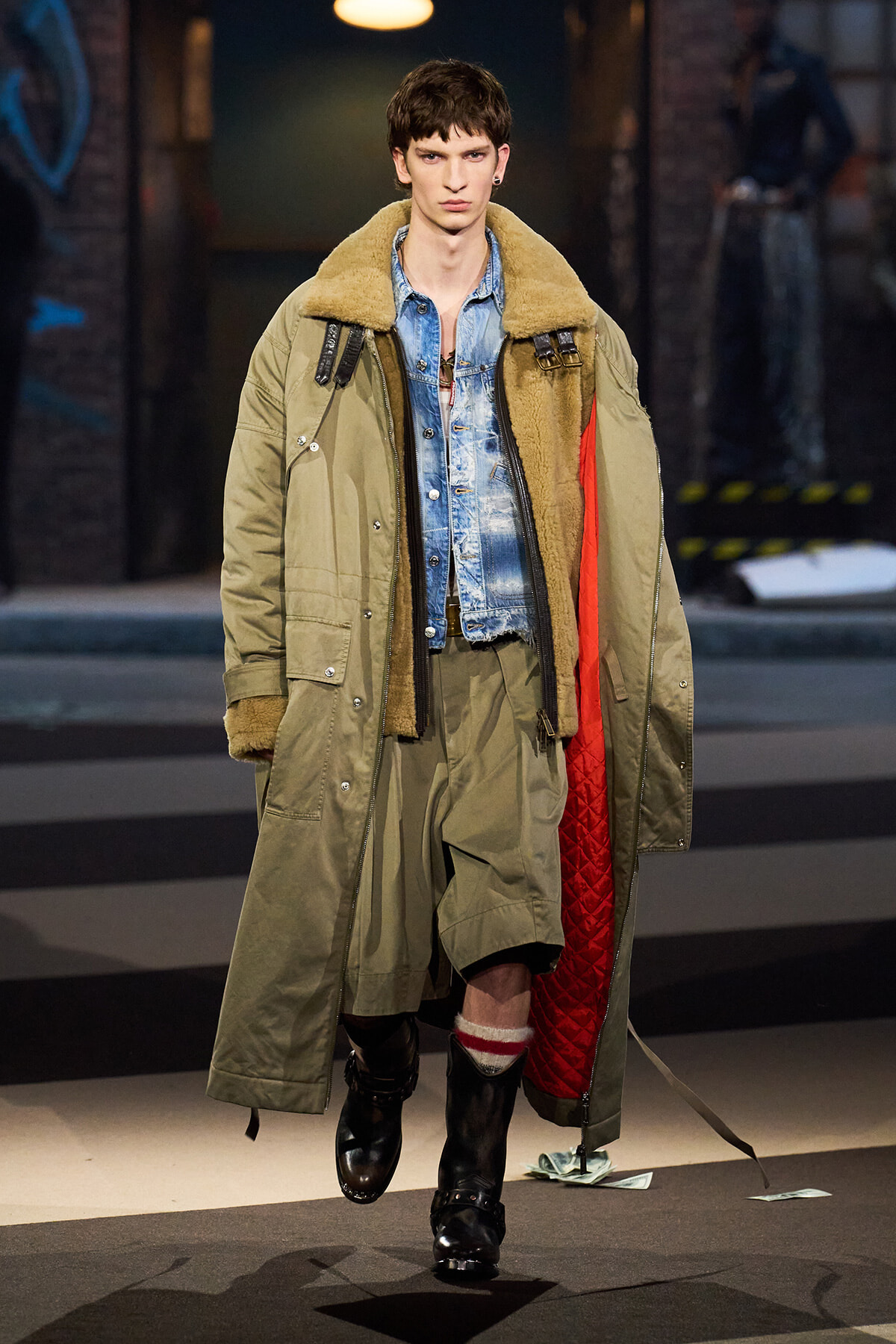 Male fashion model on the runway wearing an oversized olive parka with shearling collar over a denim jacket and baggy pants, black boots, red quilted lining visible on the inside.