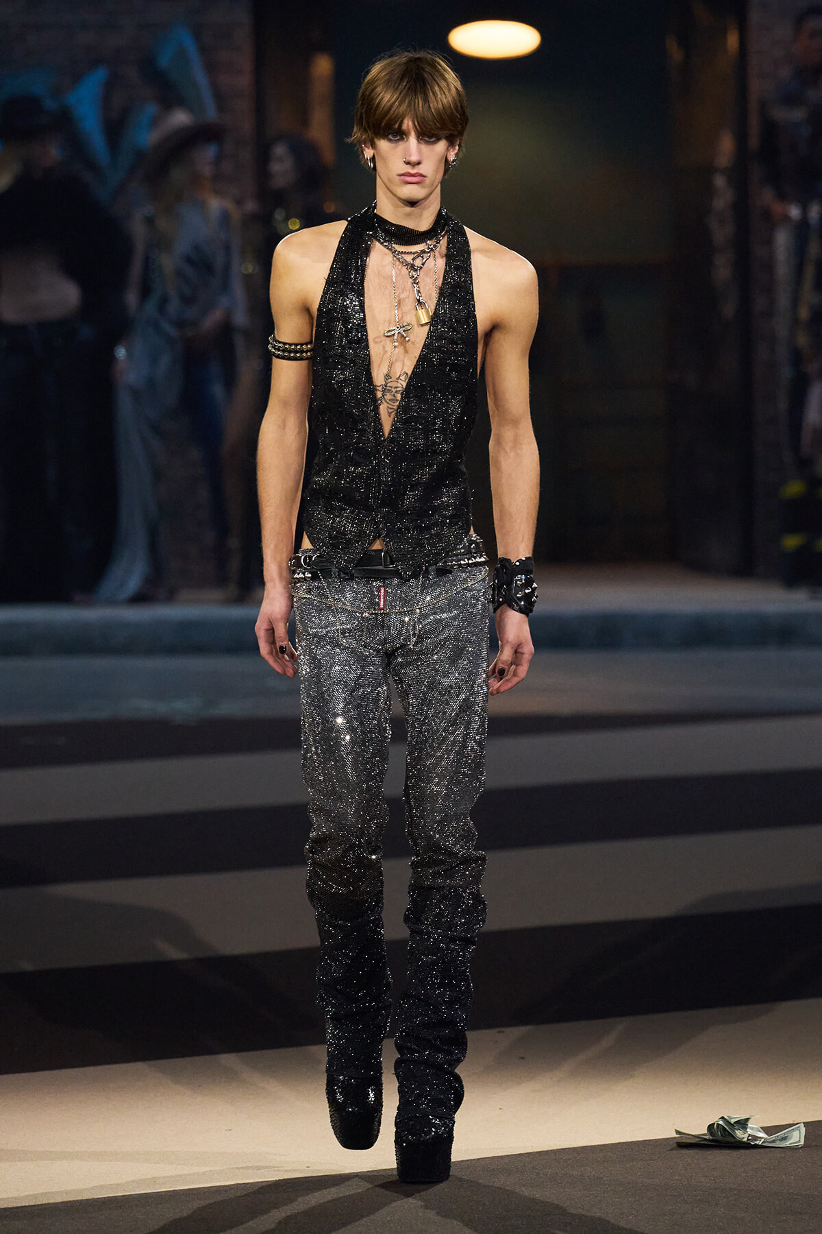 Male model on a runway wearing a black glitter halter top with a deep plunge, silver glitter pants, and layered jewelry.