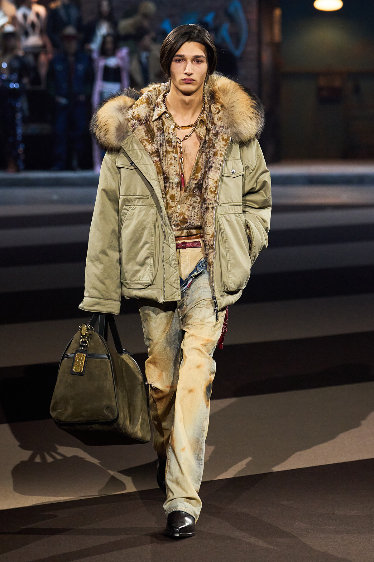 Male model walks a runway in an oversized olive parka with a fur-trimmed hood, patterned shirt, distressed jeans, and a large olive duffel bag.