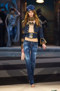 Female model walking a fashion runway in distressed denim jeans and cropped top, layered with a wide belt and denim jacket, accessorized with bold jewelry and a blue headwrap.