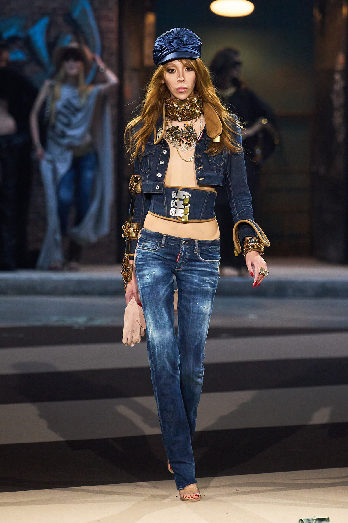Female model walking a fashion runway in distressed denim jeans and cropped top, layered with a wide belt and denim jacket, accessorized with bold jewelry and a blue headwrap.