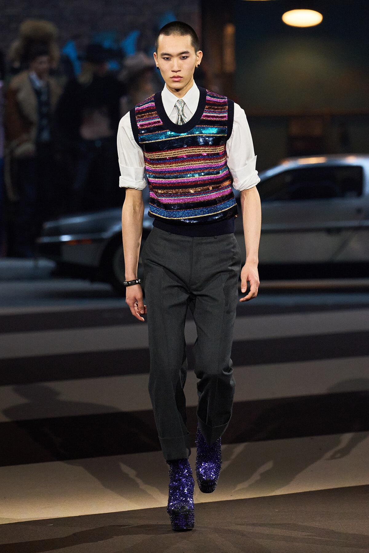Male model on a runway wearing a multicolored sequined knit vest over a white shirt, dark trousers, and glittery purple boots.