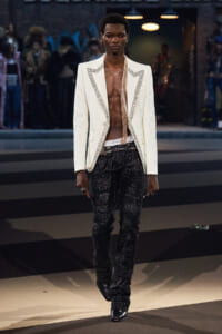 Male model walks a runway in a white studded blazer with an open chest, black glitter pants, and black shoes.
