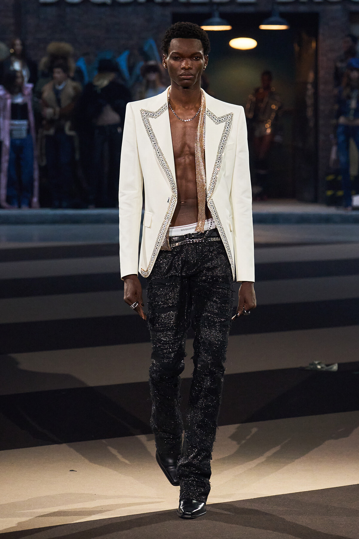 Male model walks a runway in a white studded blazer with an open chest, black glitter pants, and black shoes.