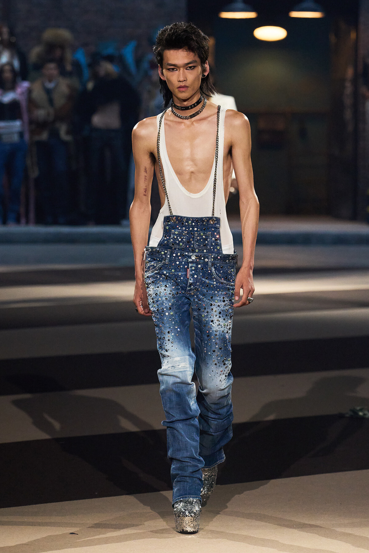 Runway model wearing a white deep-neck tank top with heavily embellished studded denim jeans and glitter platform boots, during a fashion show.