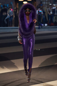 Model walking runway in a sparkling purple sequin jumpsuit with a large purple fur hood and oversized sunglasses, gloves and silver heels.