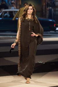 Model walks a runway in a dark brown, flowing kaftan with gold beaded trimming and oversized sleeves, complemented by gold jewelry and sandals.