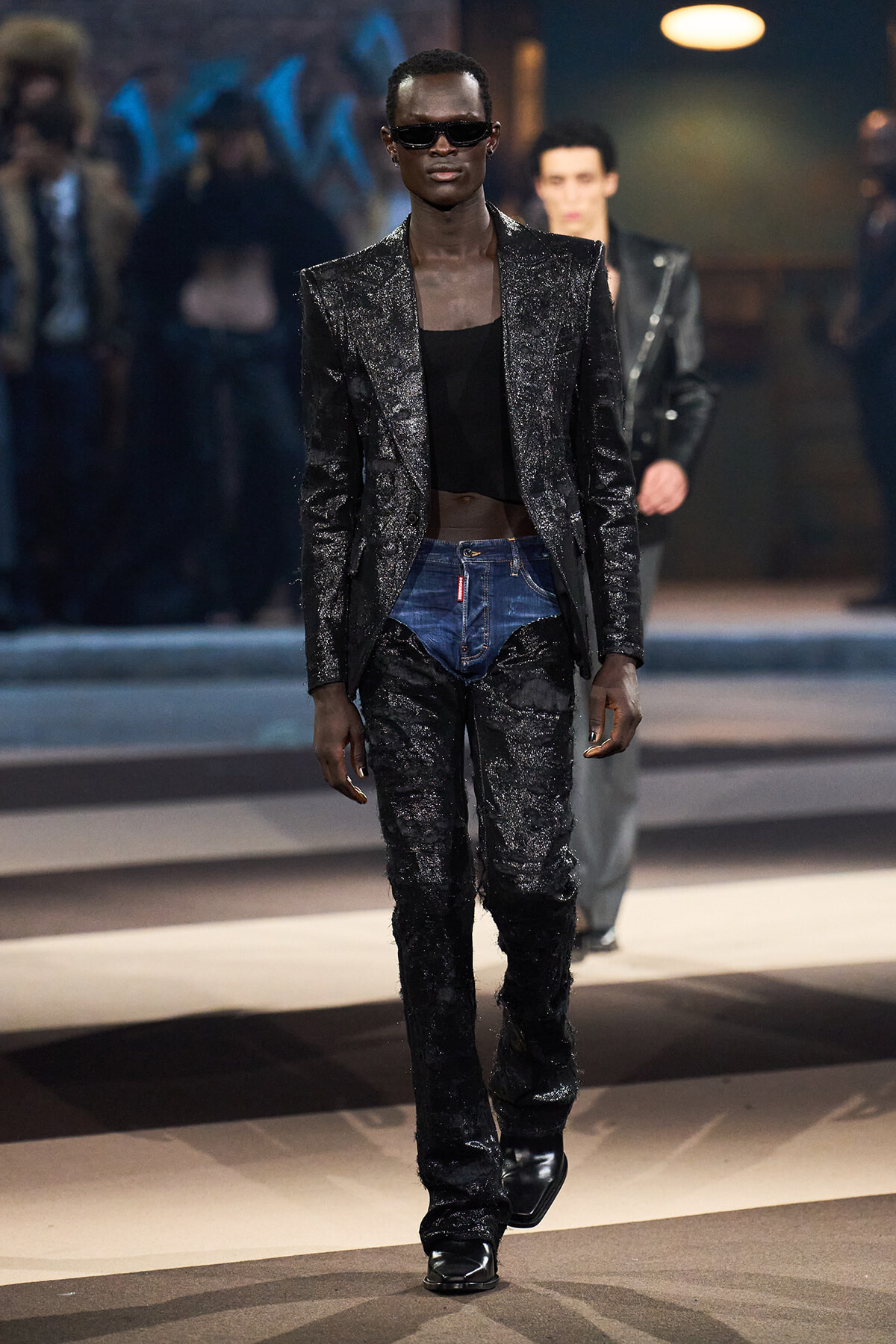 Male model walks a fashion show in a glittery black suit with a black top and jeans, wearing dark sunglasses.