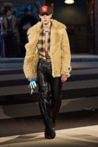 Male model walking runway in shearling tan coat over plaid shirt and black leather pants; blue and white gloves, brown cap.