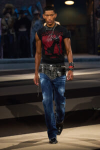 Male fashion model walking the runway in a black graphic tee, layered belts, and distressed blue jeans with boots.