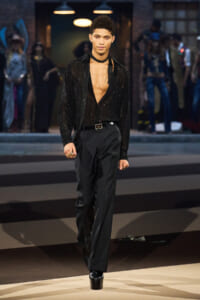 Male model walks a runway in a black sequined shirt worn open at the chest, paired with black trousers and platform boots.