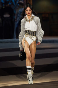 Model walks the runway in a white shirt bodysuit, oversized gray cardigan, and a wide gold‑chain belt with matching jewelry, carrying a black handbag and wearing white sneakers with striped socks.