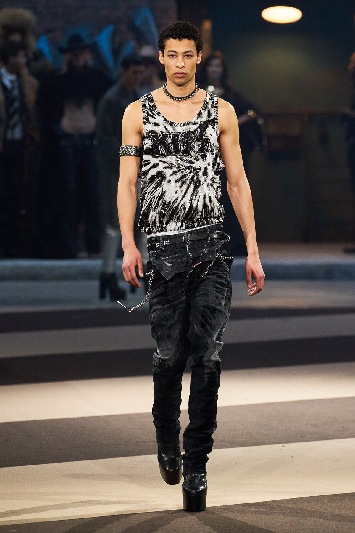 Male model walking a fashion runway in a black-and-white tie-dye sleeveless top with metal arm cuff and chain belt, black jeans, and platform boots.