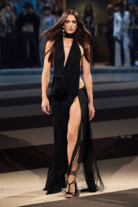 Model walking a runway in a sleeveless black gown with a deep V neckline and a high slit, fringe details at the waist, and strappy heels.
