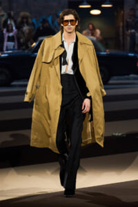 Male model walking a fashion runway in a tan trench coat over a white shirt and black waistcoat and pants, wearing oversized orange sunglasses.