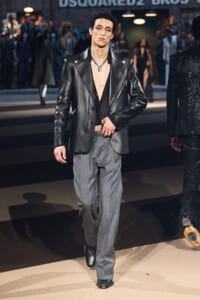 Male model walking a fashion runway in a black leather blazer, open shirt, and gray trousers with a chain necklace.