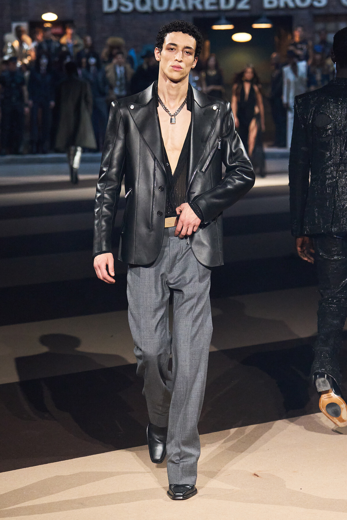 Male model walking a fashion runway in a black leather blazer, open shirt, and gray trousers with a chain necklace.