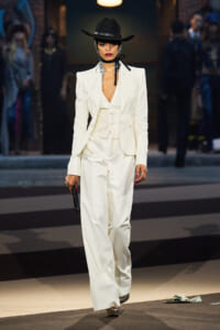 Model struts a white pantsuit with a vest and blazer on a runway, wearing a black hat and silver accessories.