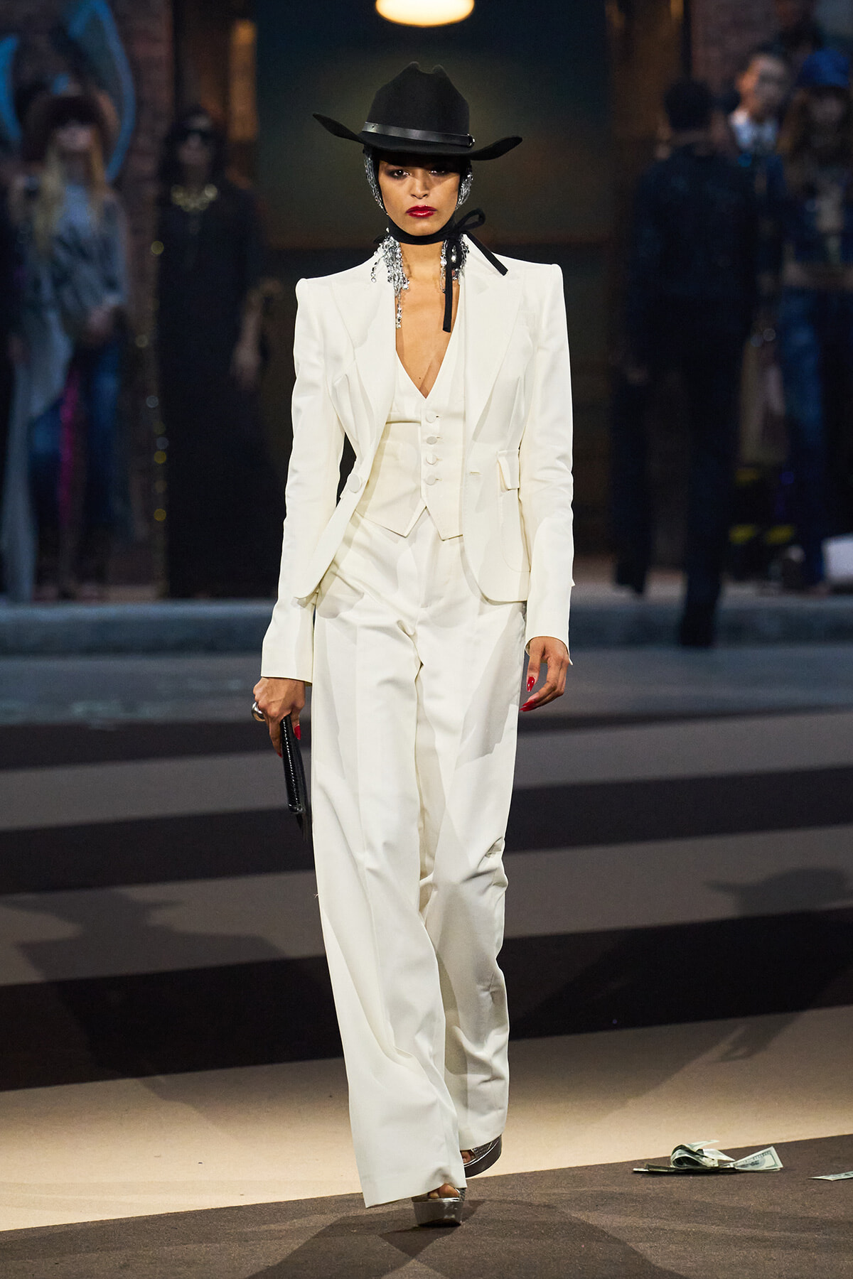 Model struts a white pantsuit with a vest and blazer on a runway, wearing a black hat and silver accessories.