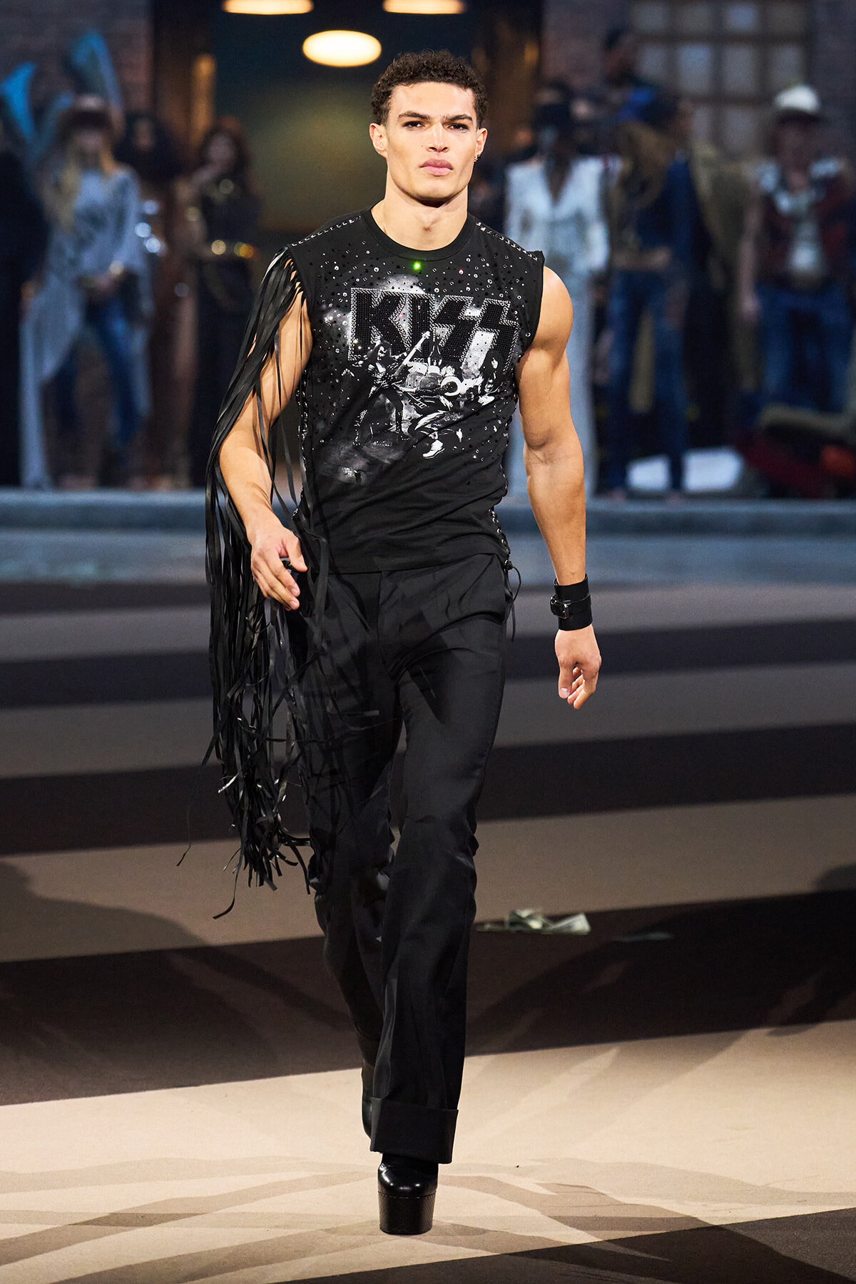 Male model walking a runway in a black fringe-sleeve top with a KISS graphic, black pants, and platform boots.