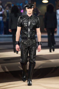 Male model on a runway wearing a black leather police-style uniform with cap and tall boots.
