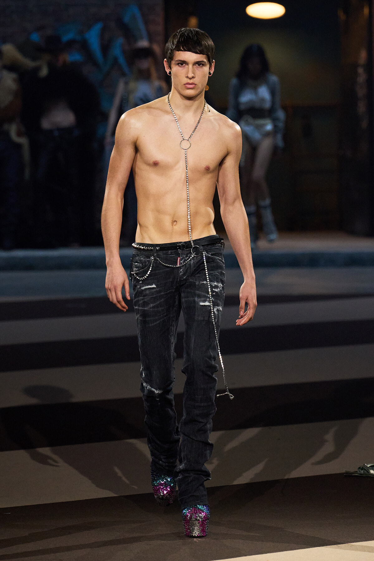 Shirtless male model walks a fashion runway wearing distressed black jeans with a long silver chain and glittery platform boots.