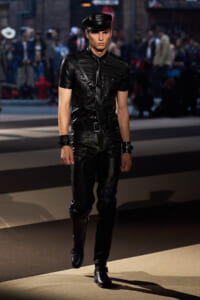 Male model in a black leather outfit with cap, belt, and crossbody strap walking a runway during a fashion show.