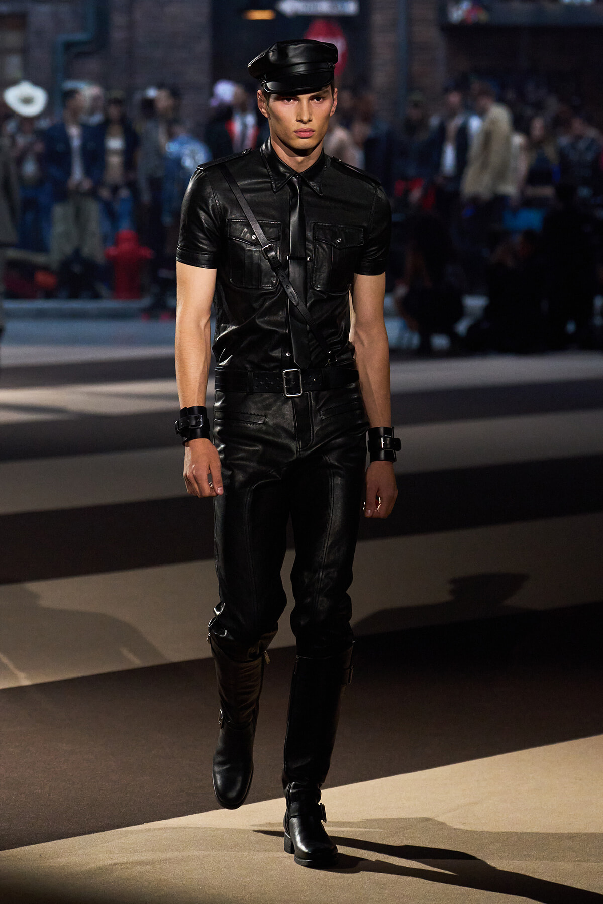 Male model in a black leather outfit with cap, belt, and crossbody strap walking a runway during a fashion show.