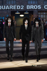 Three men in gray suits wearing Kiss-style white makeup with black accents walk a fashion runway, black platform boots visible, brick backdrop behind them—centered and dramatic.