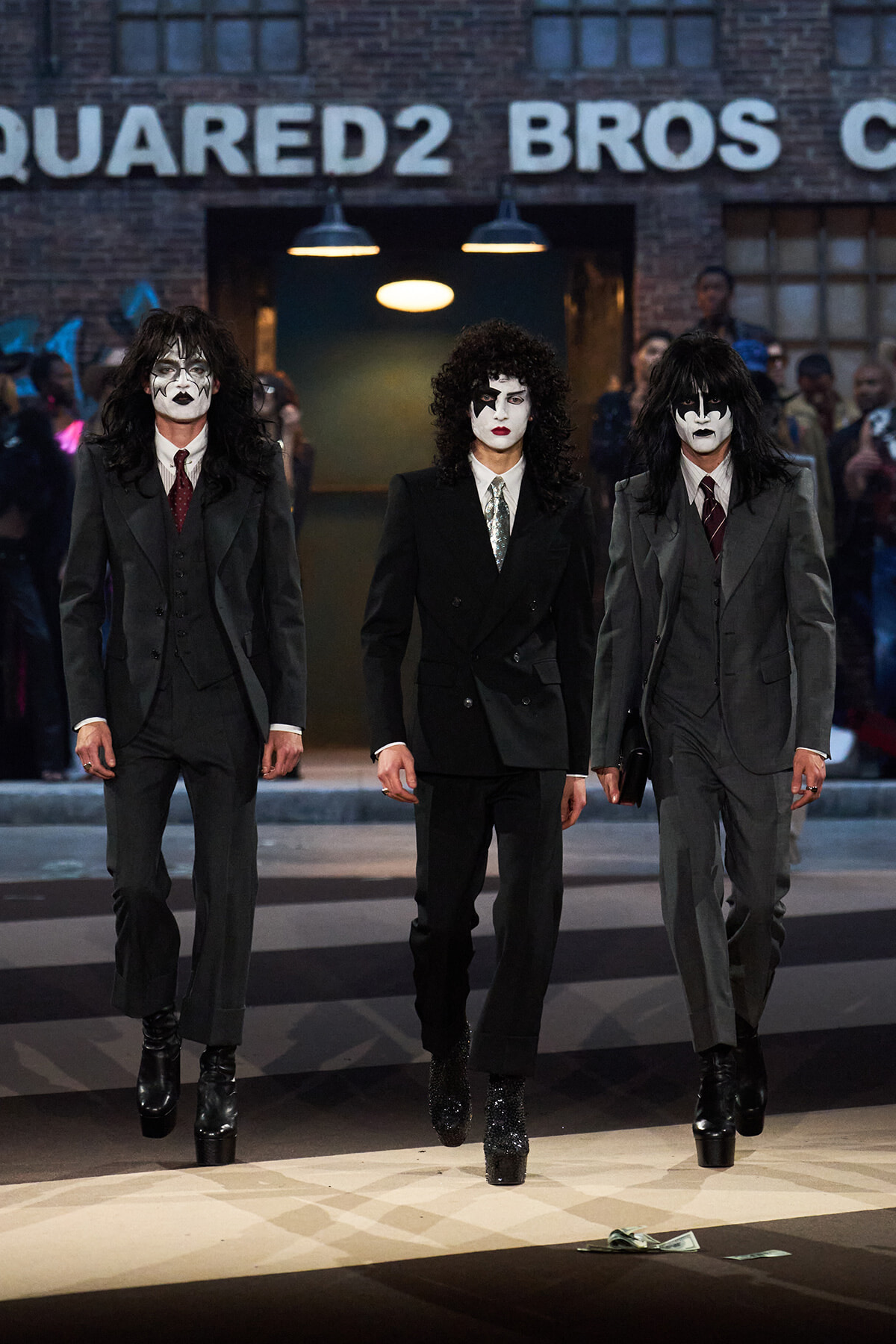 Three men in gray suits wearing Kiss-style white makeup with black accents walk a fashion runway, black platform boots visible, brick backdrop behind them—centered and dramatic.