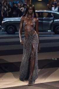 Model on a runway wearing a sparkling silver rhinestone two-piece outfit with a high-slit skirt and crisscross straps, large dangling earrings, as a vintage car and audience loom in the background.