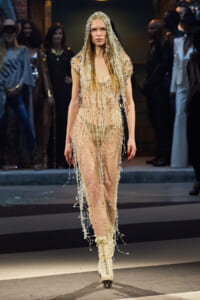 Model on the runway wearing a sheer nude beaded jumpsuit with a crystal fringe hooded overlay and cream lace-up boots with blades visible beneath the hem of the suit, as the audience watches.