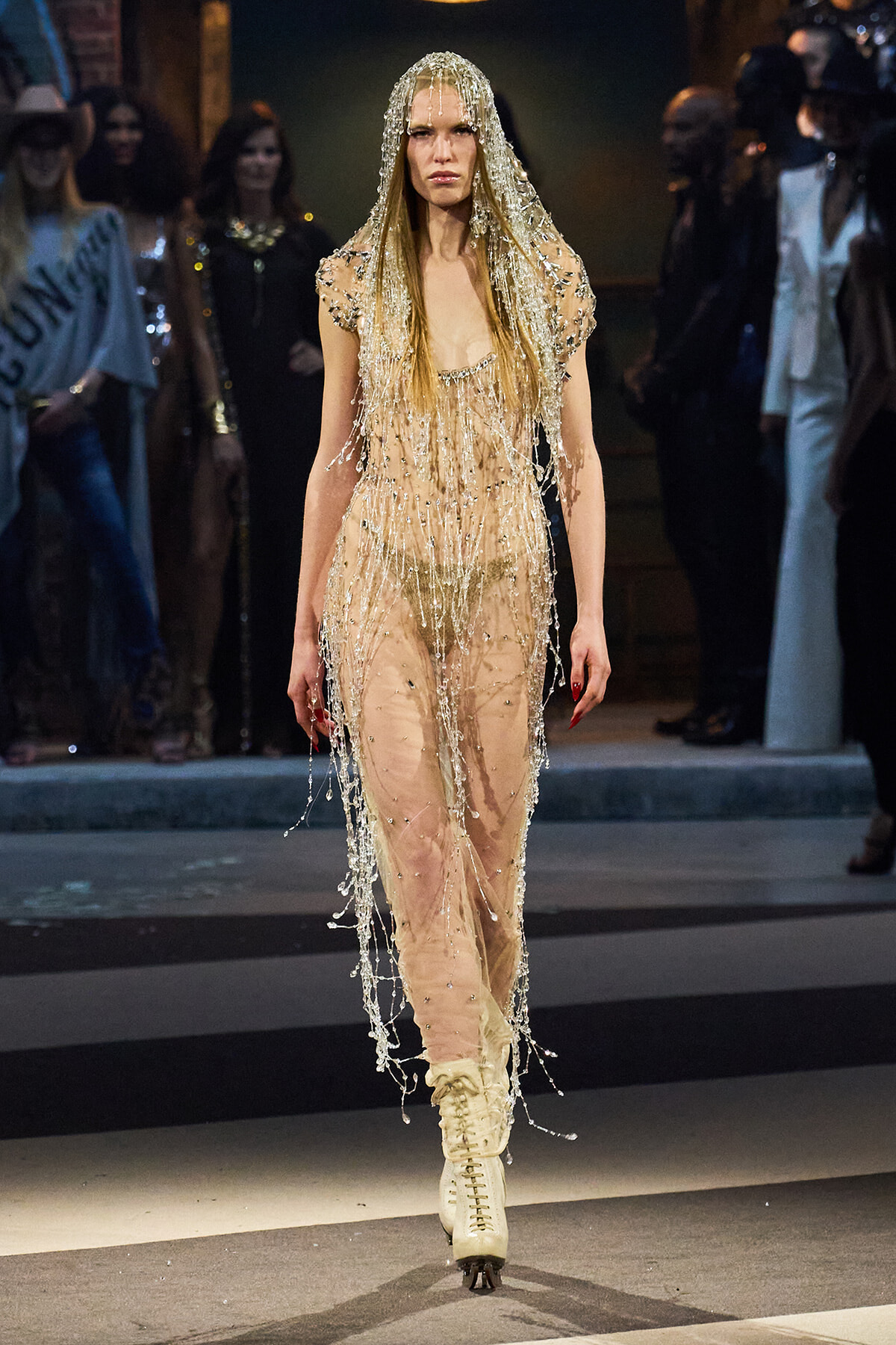 Model on the runway wearing a sheer nude beaded jumpsuit with a crystal fringe hooded overlay and cream lace-up boots with blades visible beneath the hem of the suit, as the audience watches.