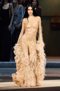 Model walking the runway in a champagne-colored sequin gown with feathered fringe sleeves and a feathered train.