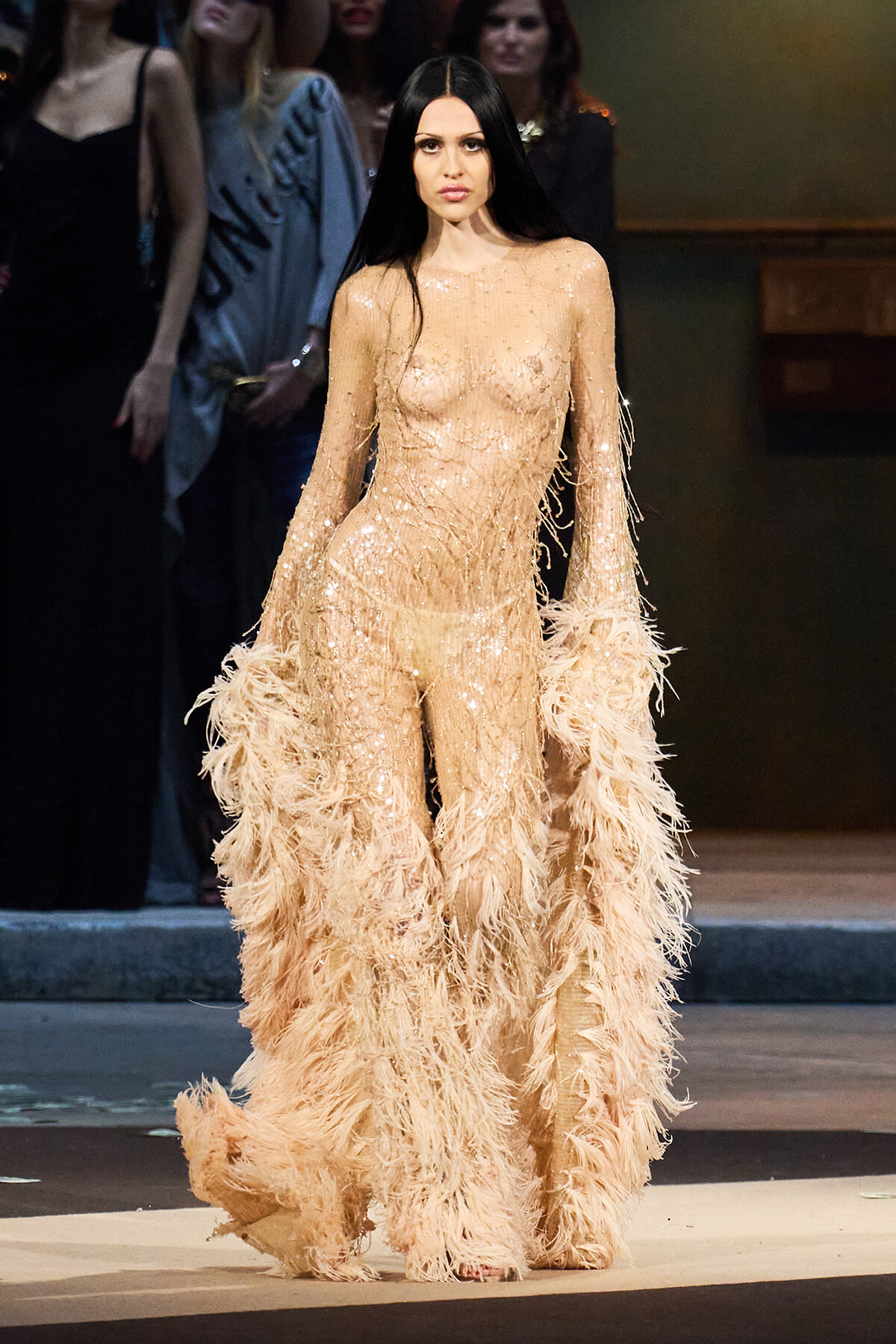 Model walking the runway in a champagne-colored sequin gown with feathered fringe sleeves and a feathered train.