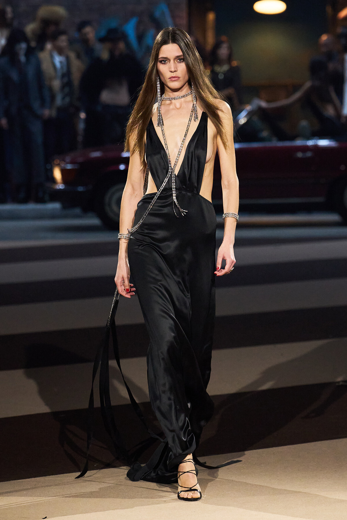 Model on a runway wearing a black satin plunging jumpsuit with long silver chain jewelry and strappy sandals.
