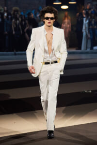 Male model in a white suit walks the fashion show runway, open polka-dot shirt and dark sunglasses.