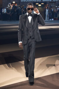 Man in a charcoal tuxedo and bow tie walks a runway, adjusting his sunglasses.