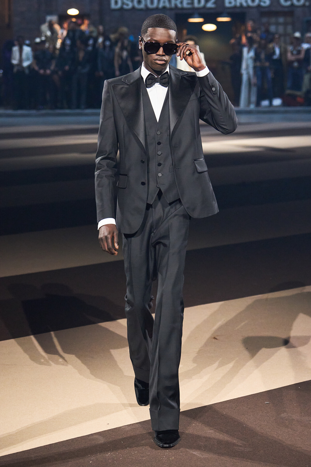 Man in a charcoal tuxedo and bow tie walks a runway, adjusting his sunglasses.