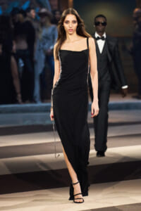 Model walks the runway in a sleek black, one-shoulder evening gown with silver chain detailing along the sides, paired with black strappy heels; a man in a tuxedo follows in the background.