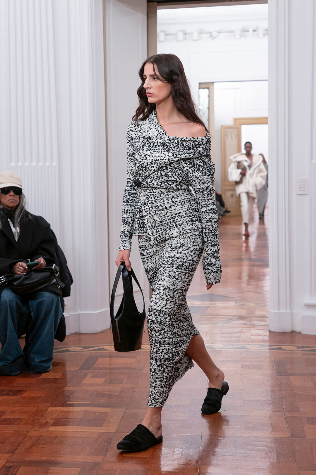 Model walks a runway in a black-and-white speckled off-the-shoulder knit dress, carrying a black handbag.
