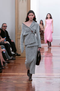 Model walking a fashion runway in a gray herringbone tweed wrap top and matching skirt, black heeled boots, red earrings, carrying a black handbag.