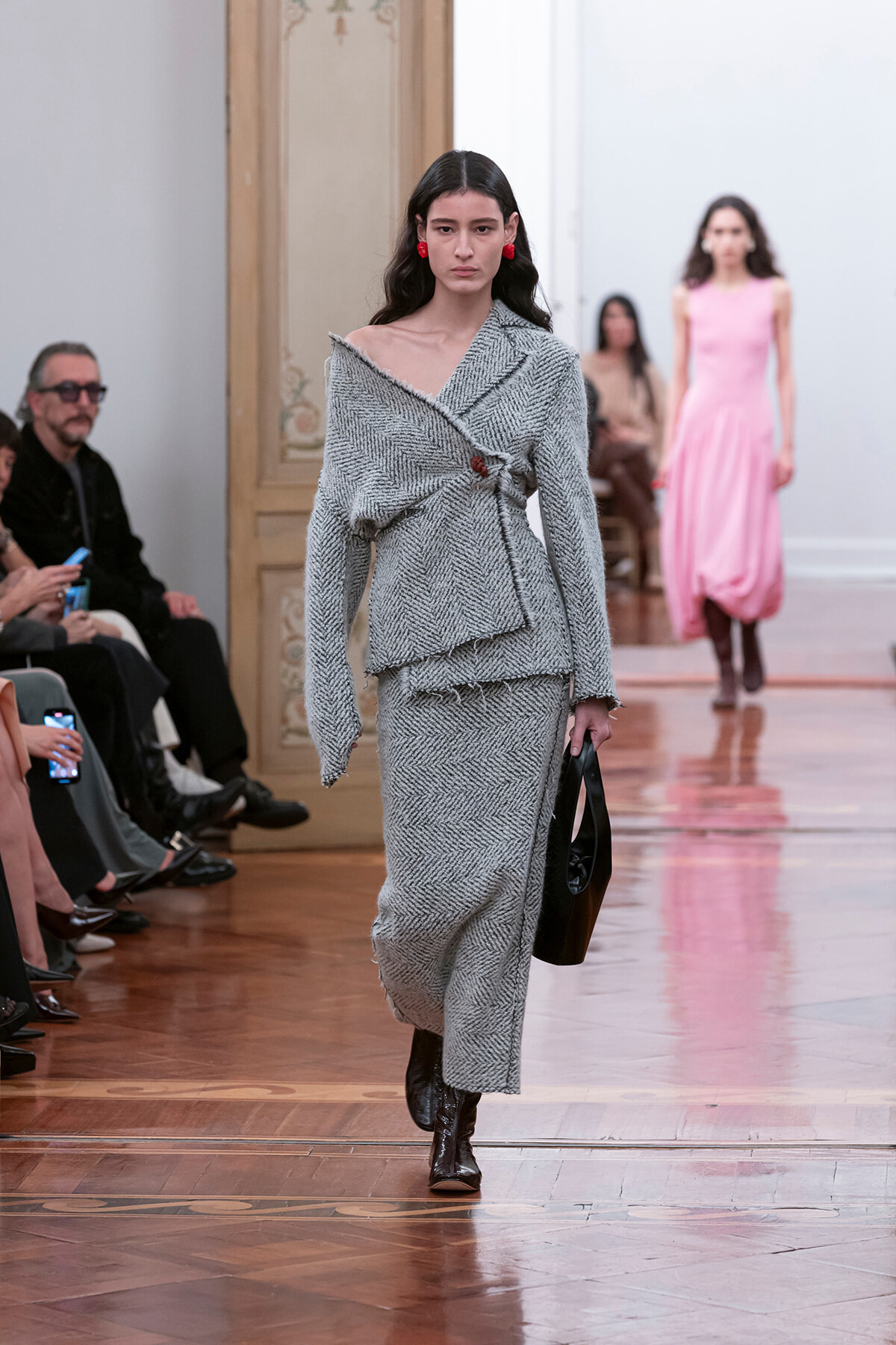 Model walking a fashion runway in a gray herringbone tweed wrap top and matching skirt, black heeled boots, red earrings, carrying a black handbag.