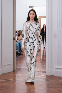 Model walking a runway in a cream satin gown with black floral vine embroidery.