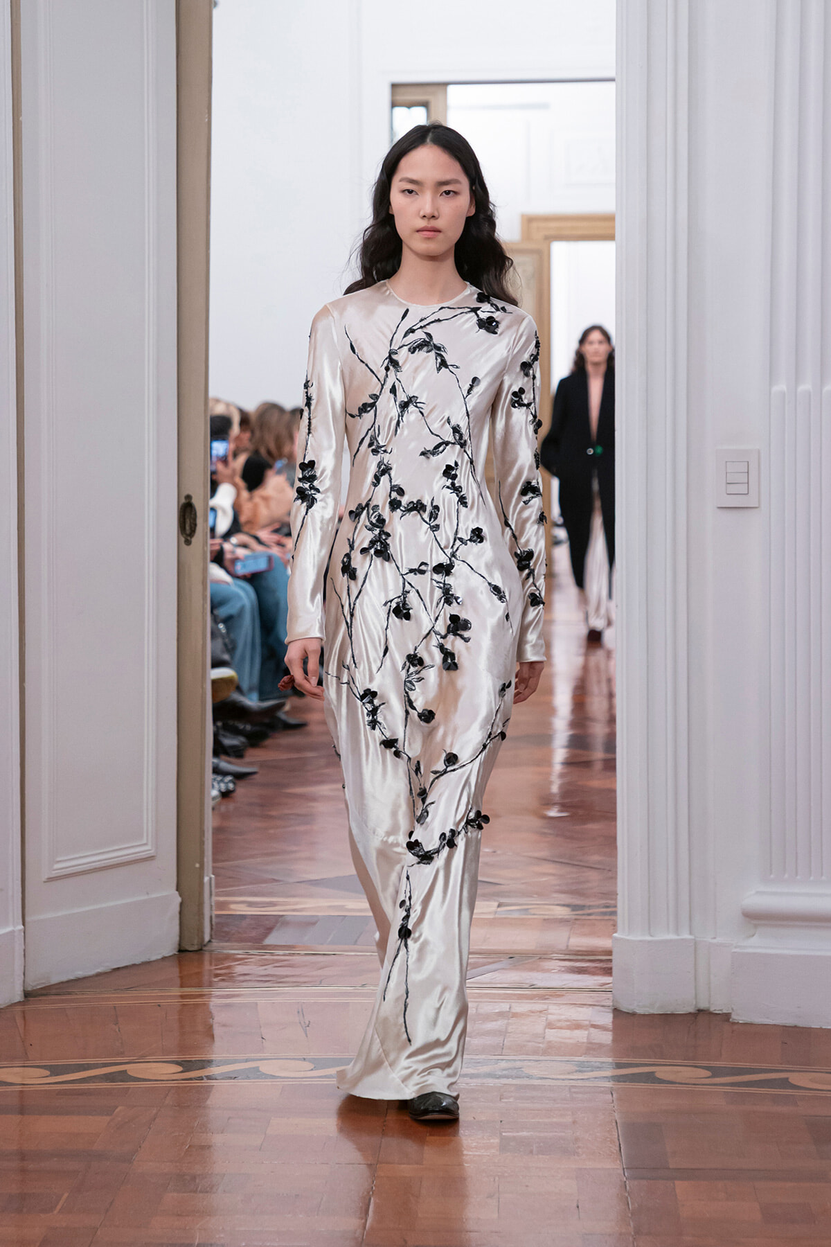 Model walking a runway in a cream satin gown with black floral vine embroidery.
