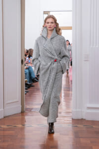 Model in a gray herringbone wrap coat walking a runway with hands in pockets and a red waist accent, black boots visible at the end of the coat. Audience watches from both sides in a bright room.